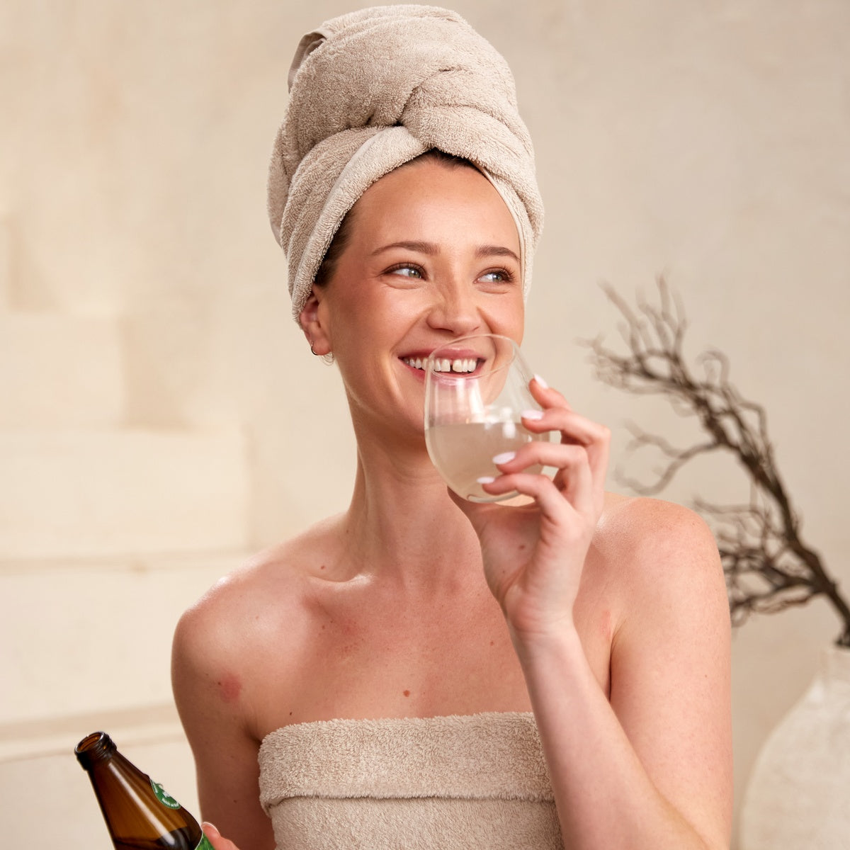 Lächelnde Frau mit Handtuch-Turban trinkt Pharmos Natur Aloe Vera BioUrsaft und hält Pharmos Natur loe Vera BioUrsaft Flasche in der Hand